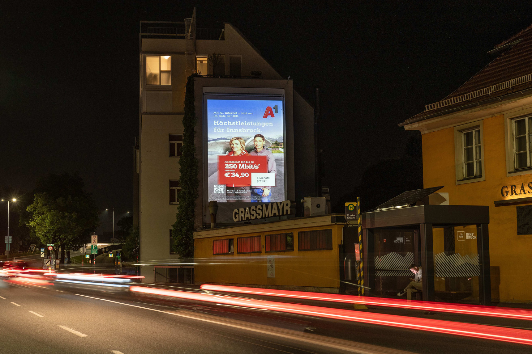 Die Hauswand eines Gebäudes neben einer Stadtstrasse wird durch eine Werbung beleuchtet in der Nacht.